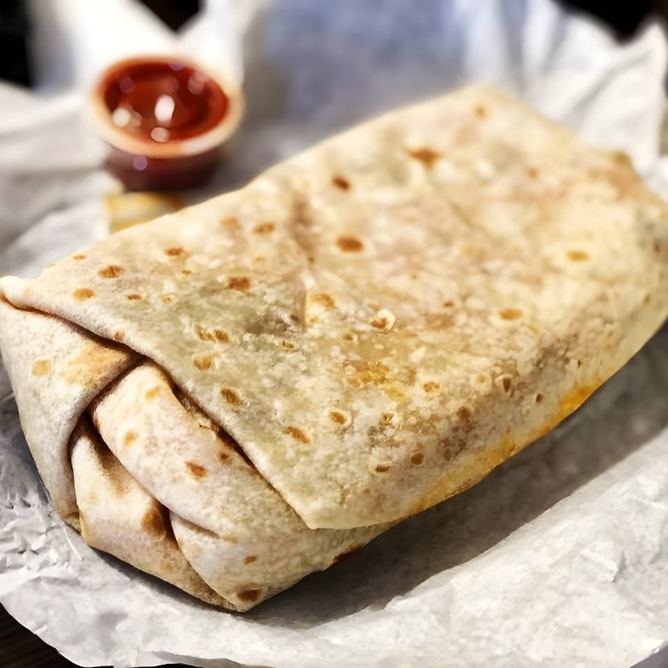 A large, warm burrito from Juanitas III, wrapped in a tortilla, with a side of red sauce.